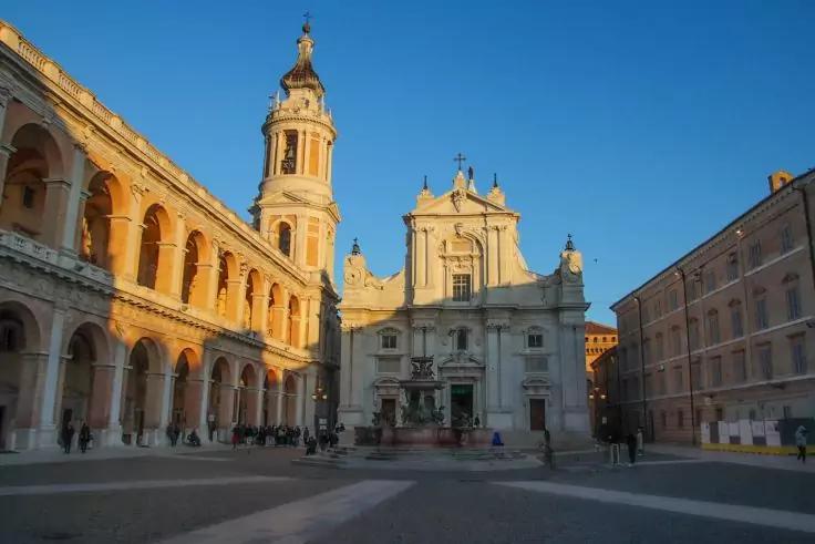 Platz mit Kirche und hohem Glockenturm links, Arkaden links, Brunnen vor der Fassade, Personen auf dem Platz, blauer Himmel