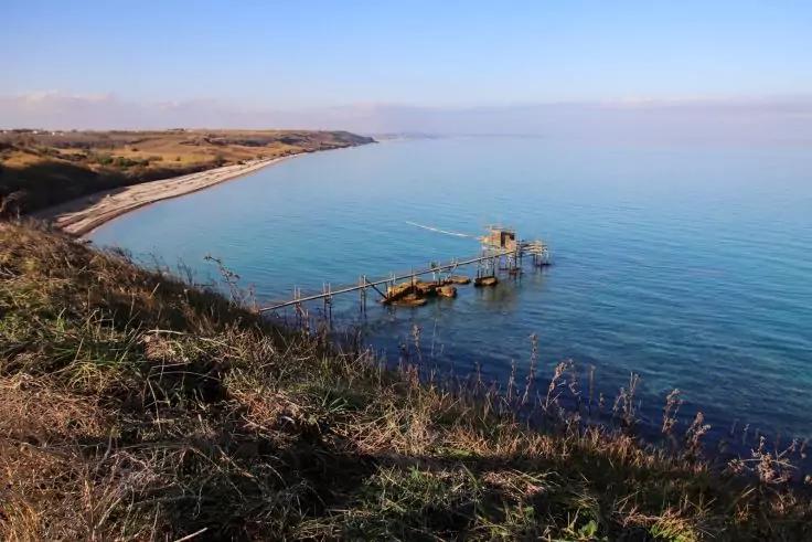 Grasbewachsener K&uuml;stenabfall im Vordergrund, darunter eine h&ouml;lzerne Plattform auf Pf&auml;hlen mit langer Stegbr&uuml;cke &uuml;ber ruhiges blaues Meer, links geschwungener Sandstrand, klarer Himmel