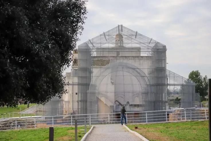 Wei&szlig;es Geb&auml;ude mit halbkreisf&ouml;rmiger Kuppel und Rundbogenfassade, vollst&auml;ndig von Ger&uuml;st und Schutznetz umgeben; im Vordergrund Weg mit Absperrung, zwei Personen stehen davor, links gro&szlig;er Baum, Grasfl&auml;chen, bew&ouml;lkter Himmel