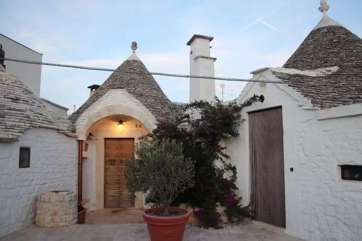 Wei&szlig;e, rundliche H&auml;user mit konischen Steind&auml;chern und Holzt&uuml;ren; gepflasterter Innenhof mit Terrakottatopf und kleinem Baum; Kletterpflanze mit rosa Bl&uuml;ten an der Fassade; blasser Abendhimmel