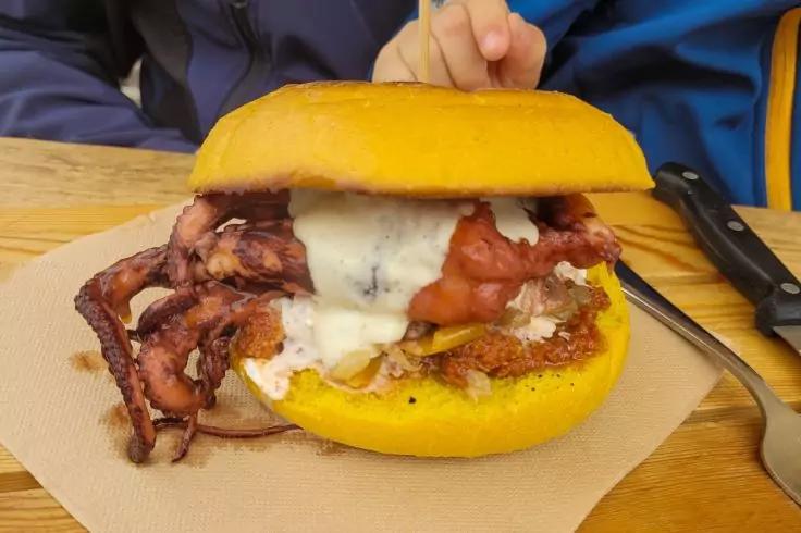 Burger mit gelbem Br&ouml;tchen, knusprigem Speck, wei&szlig;er Sauce und weiteren Zutaten auf einer Papierserviette, Gabel und Messer, Person mit blauer Jacke im Hintergrund