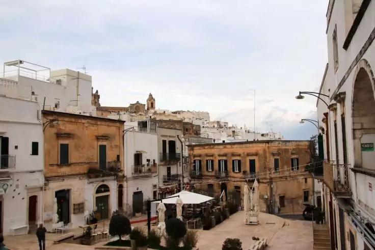 Stadtplatz mit Statue auf Sockel, umgeben von wei&szlig;en und beigen mehrst&ouml;ckigen Geb&auml;uden mit Balkonen, Tischen unter Sonnenschirm, Topfpflanzen und bew&ouml;lktem Himmel