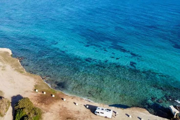 Luftaufnahme einer K&uuml;stenlinie mit t&uuml;rkisblauem Meer, sandigem Ufer, einem geparkten wei&szlig;en Wohnmobil und mehreren kleinen wei&szlig;en Gegenst&auml;nden am Strand
