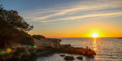 Sonne knapp &uuml;ber dem Meereshorizont, orangefarbener Himmel mit d&uuml;nnen Wolken, felsige K&uuml;ste links mit B&auml;umen, Wasser mit Lichtreflexionen