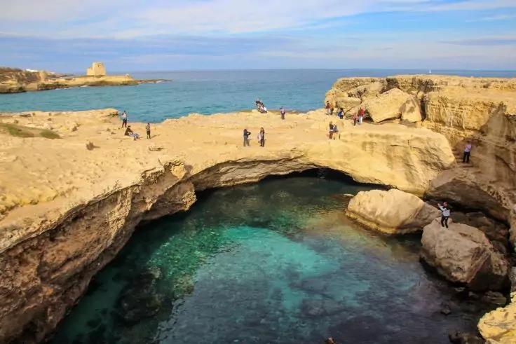 Felsbogen &uuml;ber t&uuml;rkisfarbenem Meer, mehrere Personen auf den Felsen, felsige K&uuml;ste und Horizont unter blauem Himmel mit wenigen Wolken