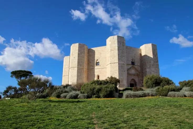 Heller steinerner achteckiger Burgbau mit runden T&uuml;rmen auf einem grasbewachsenen H&uuml;gel, davor Str&auml;ucher und ein einzelner Baum, blauer Himmel mit Wolken
