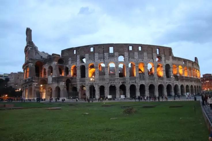 Beleuchtetes Kolosseum bei D&auml;mmerung, gr&uuml;ner Rasen im Vordergrund, vereinzelte Personen auf dem Platz