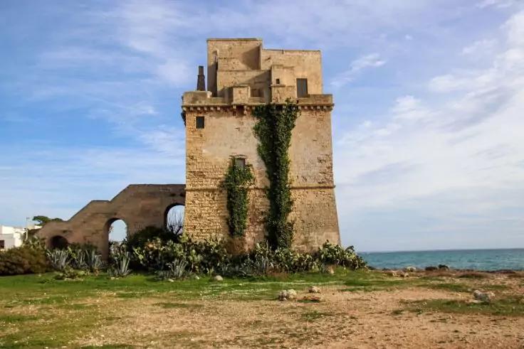 Steinerner Turm mit Kletterpflanzen, links gew&ouml;lbte Mauer, Vordergrund mit Gras und stacheligen Pflanzen, rechts Meer, blauer Himmel mit Wolken
