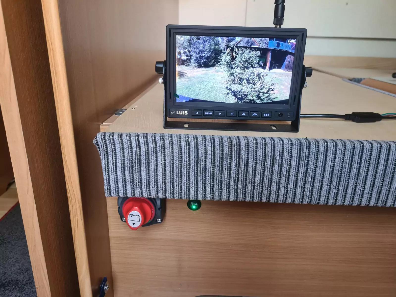 Kleiner Monitor auf einer Holzfläche; Bildschirm zeigt Garten mit Rasen, Strauch und Gebäude; grauer gerippter Stoff an der Vorderkante; roter runder Knopf und kleines grünes Licht darunter; Kabel angeschlossen