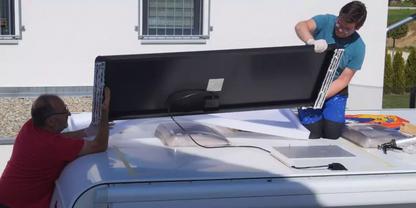 Zwei M&auml;nner heben ein schwarzes rechteckiges Paneel auf das wei&szlig;e Dach eines Vans