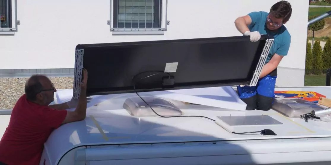 Zwei M&auml;nner heben ein schwarzes rechteckiges Paneel auf das wei&szlig;e Dach eines Vans