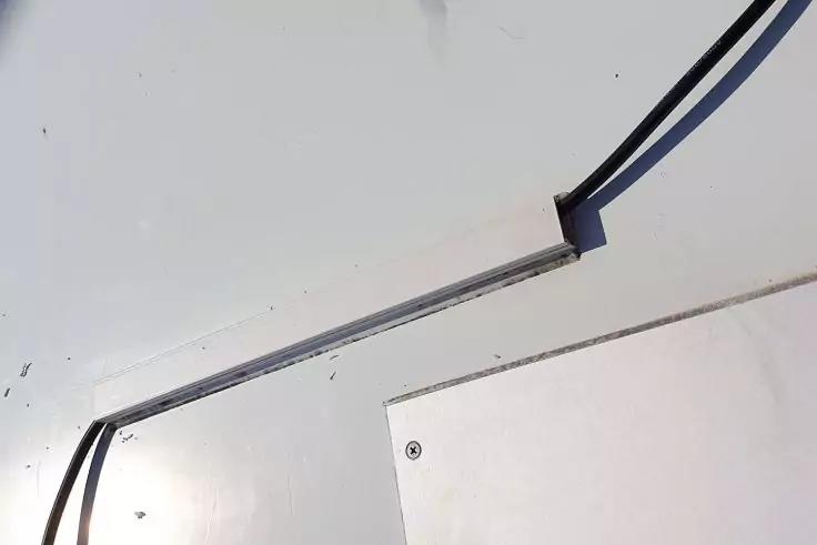 Schmales, längliches Stabgehäuse mit transparentem Streifen, zwei schwarze Kabel an den Enden, montiert auf heller Fläche; rechteckige Platte mit sichtbarer Schraube im Vordergrund