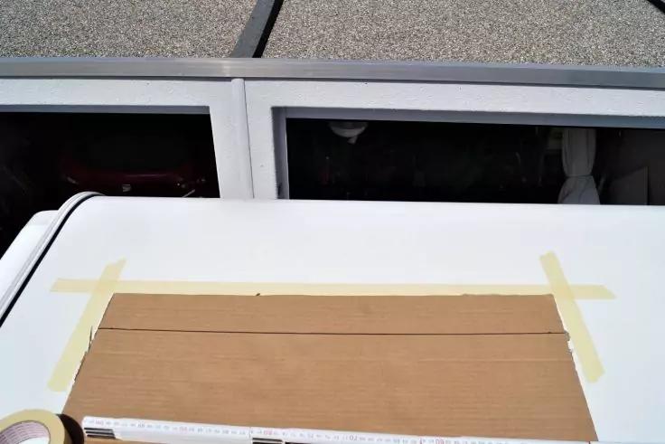 Weiße flache Oberfläche mit einem braunen Kartonstück, an den Seiten mit gelbem Klebeband befestigt; links eine Klebebandrolle, vorne ein Lineal; im Hintergrund zwei rechteckige Öffnungen über einer kiesbedeckten Fläche