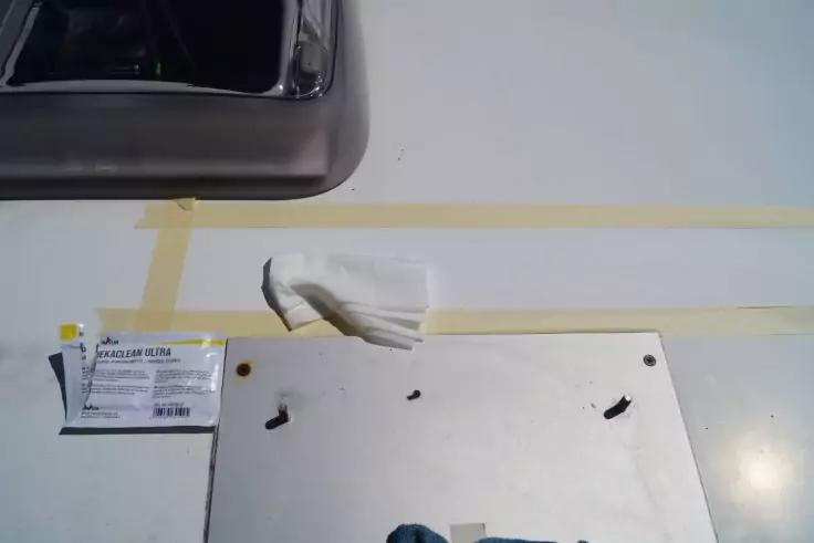 Weiße Arbeitsfläche mit gelbem Klebeband in Rechteckform, gefaltetes weißes Papiertuch in der Mitte, Edelstahlspüle oben links, Etikett links, weiße Platte mit zwei Schrauben und blauem Tuch unten