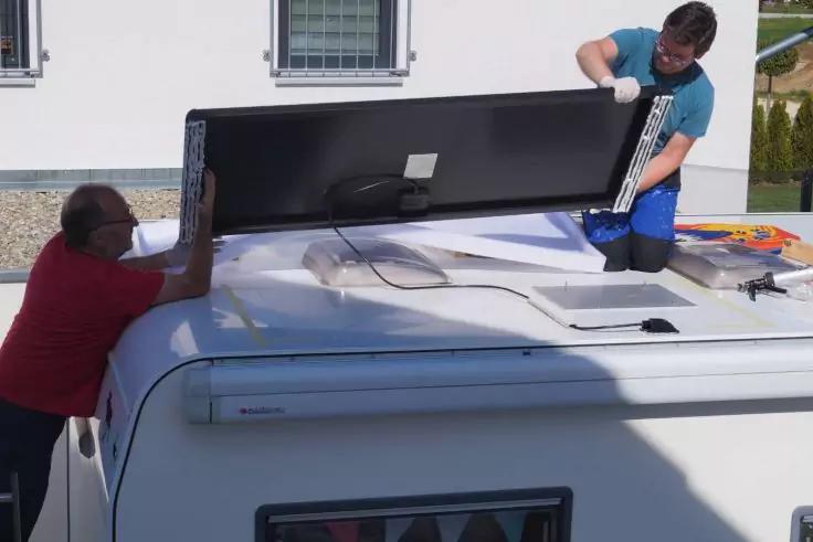 Zwei Personen auf dem Dach eines weißen Wohnmobils heben ein rechteckiges Solarpanel über eine Dachöffnung; Kabel und Werkzeuge sind sichtbar