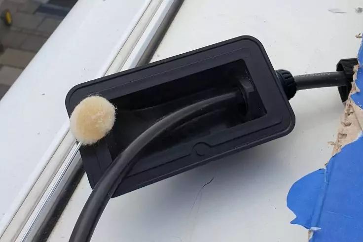 Rechteckige schwarze Kabeldurchführung in einer weißen Fläche, zwei schwarze Kabel führen hindurch, an einem Kabel eine beige Plüschkugel, rechts blaues Klebeband