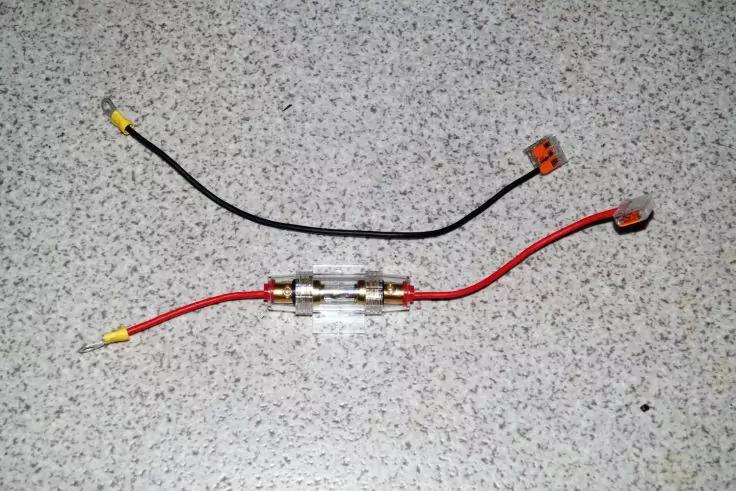 Zwei Kabel (rot und schwarz) auf grauem, gesprenkeltem Untergrund; rotes Kabel mit durchsichtiger zylindrischer Glasröhre mit Metallkappen und gelben isolierten Kabelschuhen; schwarzes Kabel mit gelben isolierten Kabelschuhen und orangefarbenem Steck