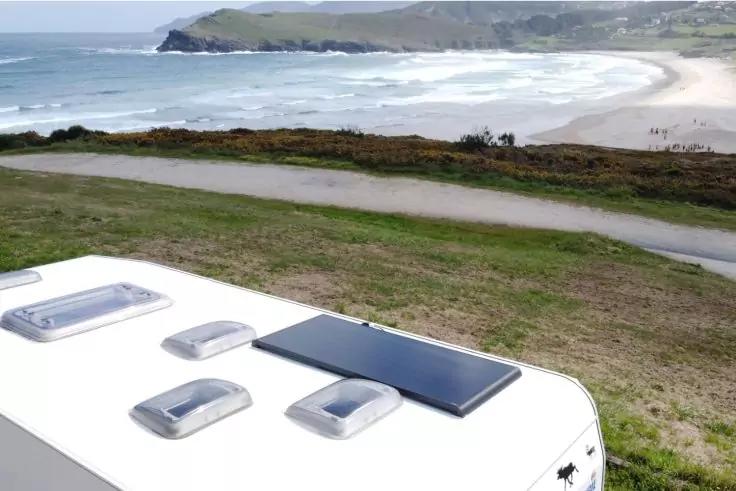 Dach eines weißen Wohnmobils mit drei Dachhauben und einem Solarpanel; im Hintergrund Gras, Weg, Sandstrand, Meer und Felsküste
