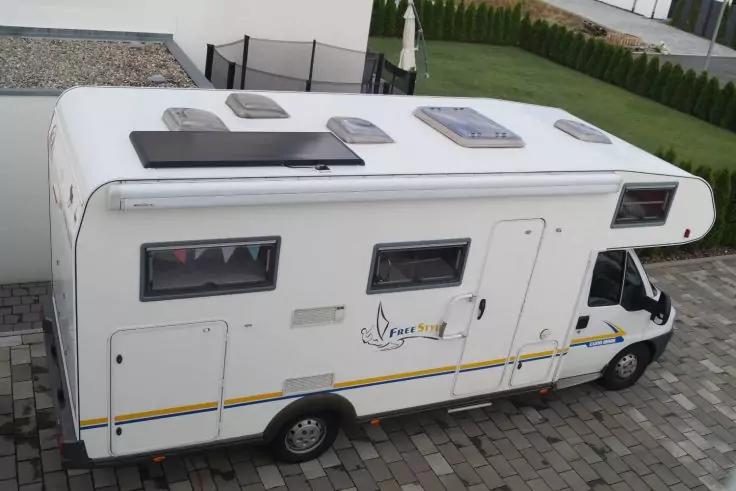 Weißes Wohnmobil mit mehreren Dachluken und einem Solarpanel auf einer gepflasterten Fläche neben einem weißen Haus und einer grünen Hecke