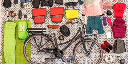 Vogelperspektive auf ein Fahrrad auf einem gemusterten Untergrund, umgeben von einem gr&uuml;nen Schlafsack, einer gr&uuml;nen Rolle, zwei roten Taschen, einer gelben Tasche, Kleidung (T-Shirts, Jacke, Shorts), einem schwarzen Helm, Schuhen, Wasserflaschen, ei