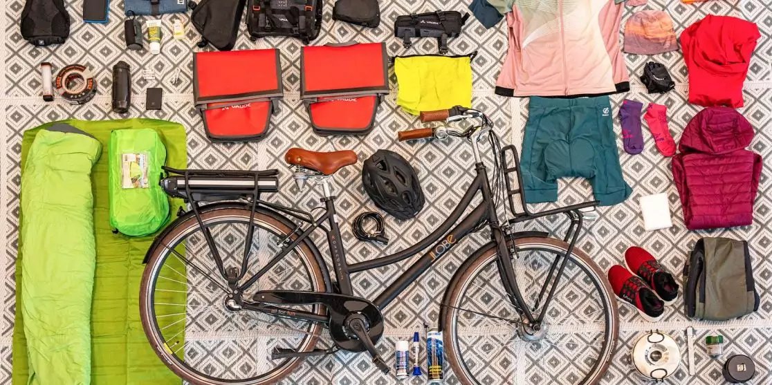 Vogelperspektive auf ein Fahrrad auf einem gemusterten Untergrund, umgeben von einem gr&uuml;nen Schlafsack, einer gr&uuml;nen Rolle, zwei roten Taschen, einer gelben Tasche, Kleidung (T-Shirts, Jacke, Shorts), einem schwarzen Helm, Schuhen, Wasserflaschen, ei