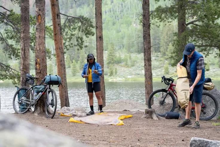 Zwei Personen neben beladenen Fahrr&auml;dern im Wald an einem See. Eine Person steht vor einer gelben Plane auf dem Boden und h&auml;lt ein gelbes Teil, die andere h&auml;lt einen zusammengerollten beigen Gegenstand. Kiefern und See im Hintergrund