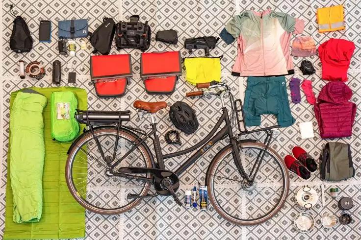 Draufsicht auf ein Fahrrad in der Mitte, umgeben von Camping- und Reiseausr&uuml;stung: gr&uuml;ner Schlafsack und Isomatte, rote und gelbe Packtaschen, Helm, Jacken, Shirts, Schuhe und Sandalen, Rucksack, Kamera, Flaschen, Werkzeug und kleine Gegenst&auml;nde