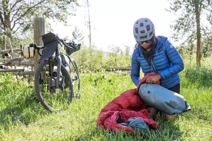 Person mit Fahrradhelm und Sonnenbrille kniet auf Gras und pumpt eine graue aufblasbare Schlafmatte; roter Schlafsack daneben, Fahrrad lehnt an Holzzaun, B&auml;ume im Hintergrund