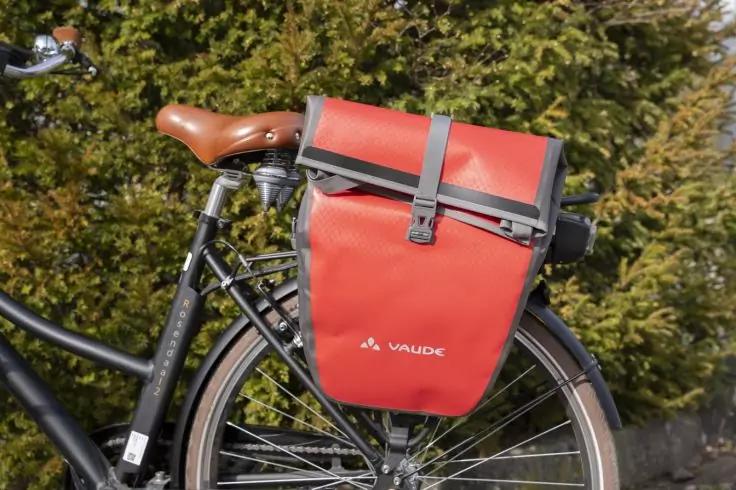R&uuml;ckteil eines Fahrrads mit braunem Ledersattel und roter Vaude-Packtasche am Gep&auml;cktr&auml;ger, gr&uuml;ne Hecke im Hintergrund