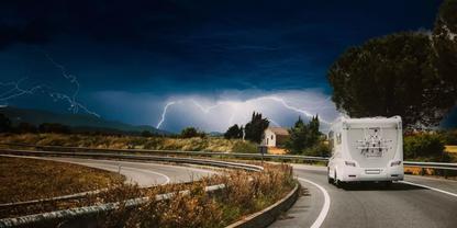 Wohnmobil auf kurviger Landstra&szlig;e, dunkle Gewitterwolken mit sichtbaren Blitzen, kleines Haus am Stra&szlig;enrand, Leitplanke und trockenes Gras