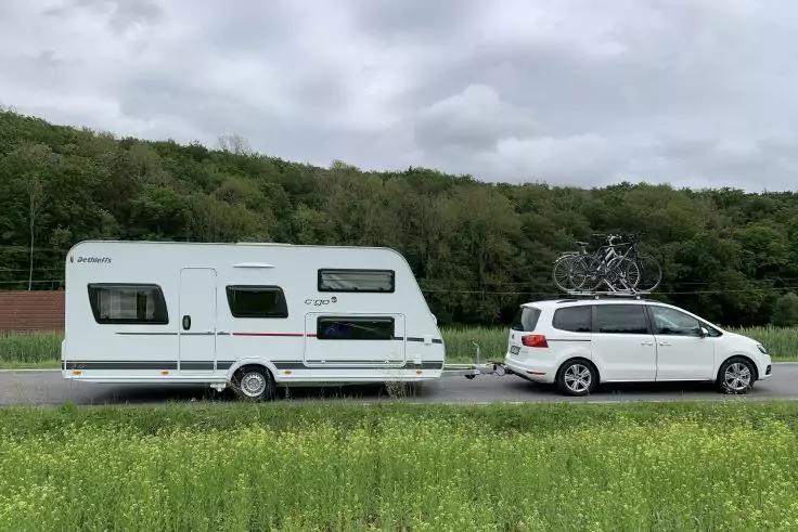 Wei&szlig;es Auto mit zwei Fahrr&auml;dern auf dem Dach zieht einen wei&szlig;en Wohnwagen auf einer Landstra&szlig;e, Wiese im Vordergrund, bewaldete H&uuml;gel und bew&ouml;lkter Himmel im Hintergrund