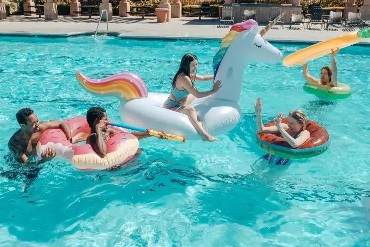 F&uuml;nf Personen auf aufblasbaren Schwimmhilfen in einem Pool: eine auf einem gro&szlig;en wei&szlig;en Einhorn, zwei auf einem pinken Donut, eine auf einem roten Donut, eine auf einem gelben Paddleboard; Liegest&uuml;hle und Sonnenschirme am Beckenrand