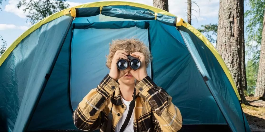 Person mit kariertem Hemd sitzt im Eingang eines blauen Zelts und schaut durch ein Fernglas; B&auml;ume im Hintergrund