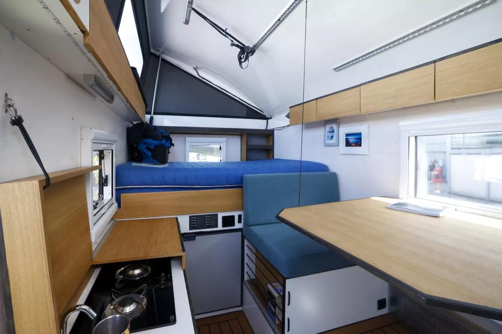 Innenraum mit blauem Sitzpolster, Holzm&ouml;beln, Tisch, Kochnische mit Herd und Sp&uuml;le, Liegefl&auml;che und Fenstern