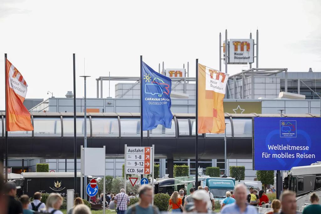 Personen und Fahrzeuge im Vordergrund vor einem Geb&auml;ude; mehrere Fahnen auf Masten, blaues Schild mit wei&szlig;er Aufschrift