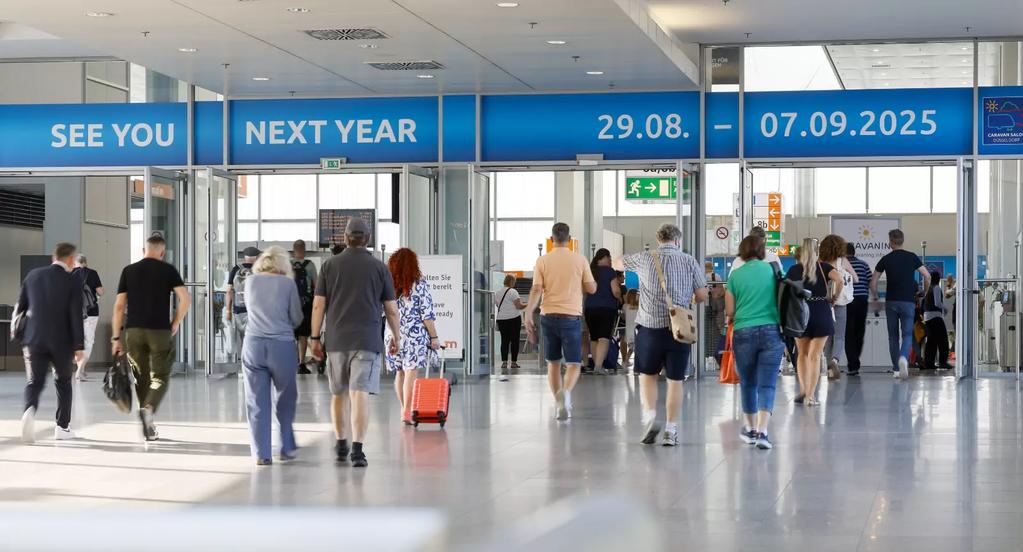 Mehrere Personen gehen in einer gro&szlig;en, hellen Halle mit Glasw&auml;nden; einige ziehen Koffer. &Uuml;ber ihnen h&auml;ngen blaue digitale Tafeln mit dem Text "SEE YOU NEXT YEAR" und den Daten "29.08. - 07.09.2025"