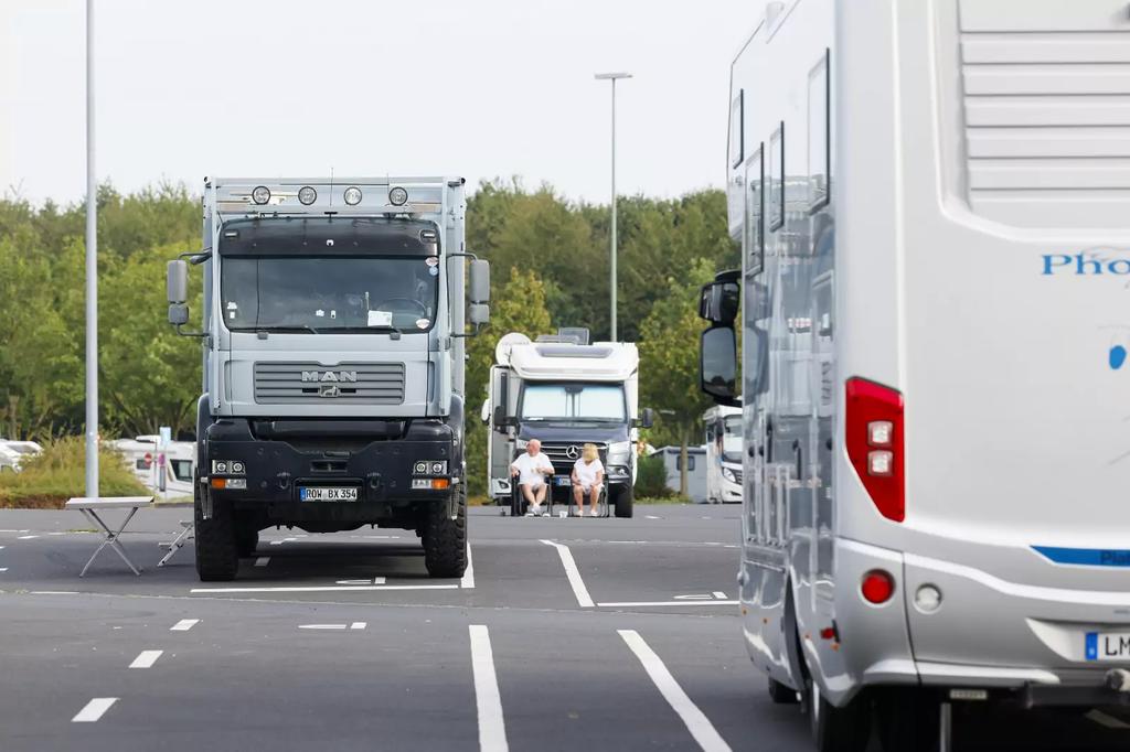 Parkplatz mit Wohnmobilen: vorn rechts die R&uuml;ckseite eines wei&szlig;en Wohnmobils, links ein Lkw, in der Mitte ein Wohnmobil und zwei Personen auf Campingst&uuml;hlen, B&auml;ume im Hintergrund