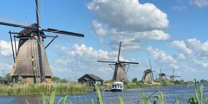 Mehrere Windm&uuml;hlen entlang eines Kanals, Wasser mit kleinem wei&szlig;en Boot, Schilf im Vordergrund, H&auml;user am Ufer, blauer Himmel mit Wolken