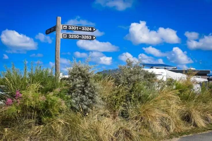 Holzwegweiser mit zwei schwarzen Richtungsschildern mit wei&szlig;en Zahlen vor hohem Gras und Str&auml;uchern, rechts teilweise sichtbares wei&szlig;es Geb&auml;ude, blauer Himmel mit vereinzelten Wolken