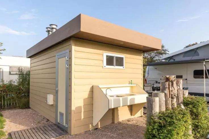 Kleines beiges Geb&auml;ude mit flachem Dach, kleines Seitenfenster, Au&szlig;ensp&uuml;le an der Wand, T&uuml;r, Holzpfosten und B&uuml;sche daneben, Wohnwagen im Hintergrund, blauer Himmel