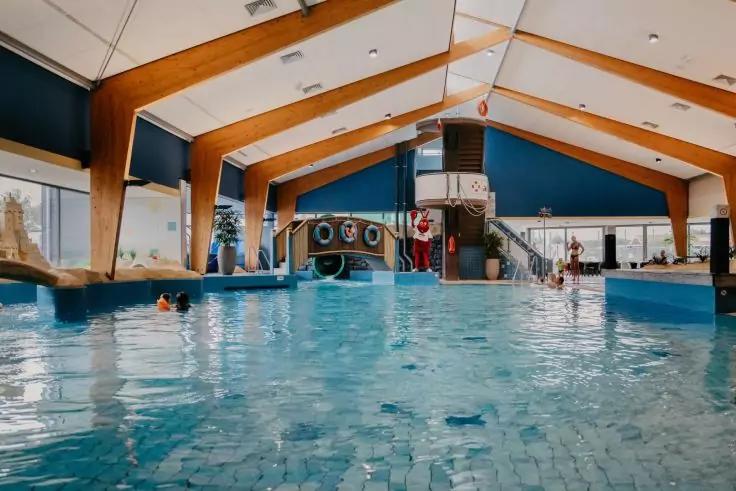 Hallenbad mit blau gefliestem Becken, sichtbaren Holztr&auml;gern an der Decke, Spielturm mit Rutsche und Rettungsring, mehrere Personen im Wasser und gro&szlig;e Fenster