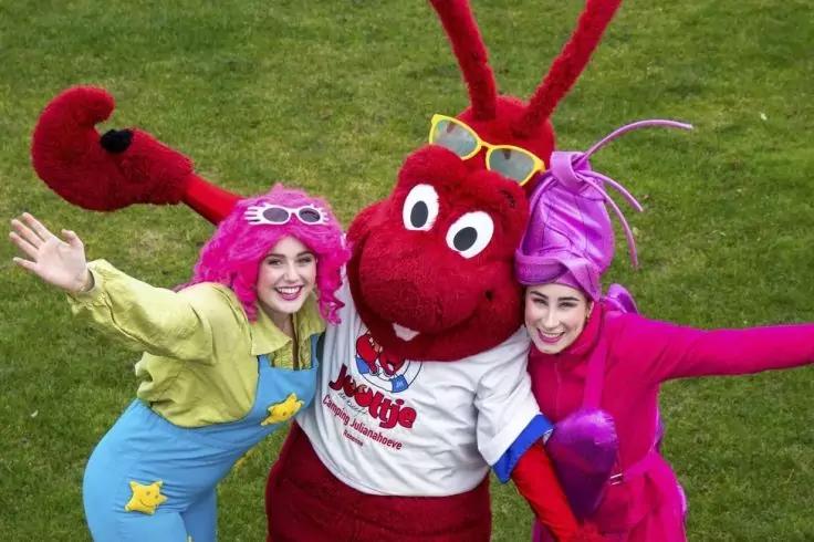 Drei verkleidete Personen auf Rasen: links gelbe Jacke, blaue Latzhose, pinke Per&uuml;cke und Brille; Mitte gro&szlig;es rotes Maskottchen mit gro&szlig;en Augen, Antennen und wei&szlig;em T‑Shirt; rechts pink‑violettes Kost&uuml;m und violette Per&uuml;cke. Alle haben die Arme aus