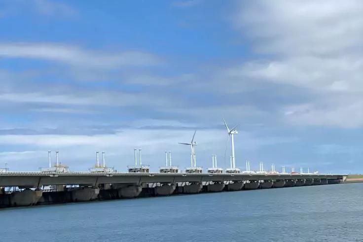 Lange Betonbr&uuml;cke mit Lampenmasten, drei Windr&auml;der im Hintergrund, Wasser im Vordergrund und wolkiger Himmel