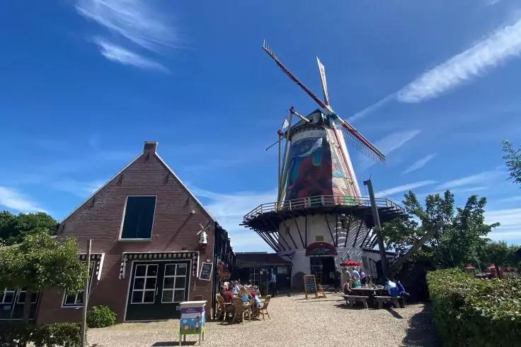 Windm&uuml;hle mit Fl&uuml;geln neben einem Backsteinhaus, blauer Himmel, Menschen, Tische auf Kiesinnenhof, B&auml;ume