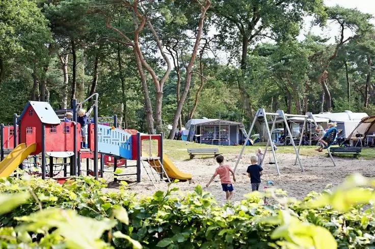 Spielplatz mit Kletterger&uuml;st, gelben Rutschen und Schaukeln; zwei Kinder laufen; Wohnwagen und Zelte im Hintergrund; B&auml;ume und Str&auml;ucher im Vordergrund