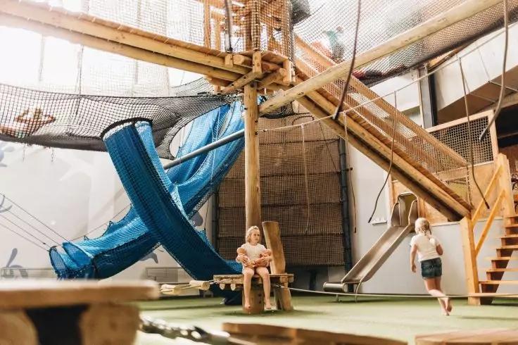 Innenbereich eines Holz-Spielger&uuml;sts mit blauem Rutschrohr, Netzen, Treppe und zwei Personen, eine sitzt auf einem Holzstuhl