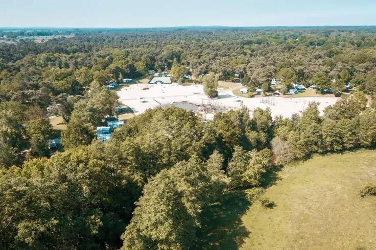 Luftaufnahme eines Waldgebiets mit einer hellen offenen Fl&auml;che, mehreren wei&szlig;en Fahrzeugen und kleinen Geb&auml;uden, im Vordergrund B&auml;ume und eine Wiese