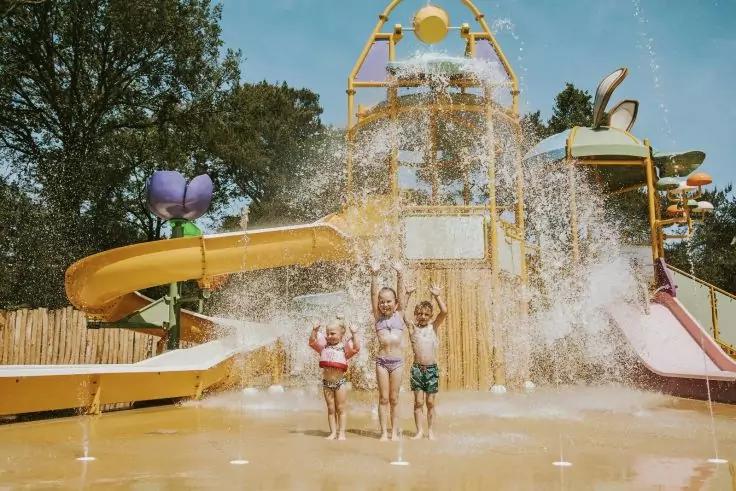 Drei Kinder stehen auf einem Wasserspielplatz, ein gro&szlig;er Wassereimer sch&uuml;ttet Wasser &uuml;ber ihnen; im Hintergrund gelbe und rosa Rutschen und B&auml;ume