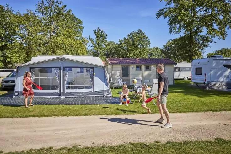 Zwei Erwachsene und zwei Kinder vor einem Wohnwagen mit angebautem Vorzelt auf einem Campingplatz; eine Person tritt einen Ball auf einem Weg