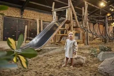 Innenraum mit Holzger&uuml;st, Metallrutsche, Seilen und sandigem Boden; barf&uuml;&szlig;ige Person in hellem Kleid steht neben Felsen; Pflanzenbl&auml;tter im Vordergrund